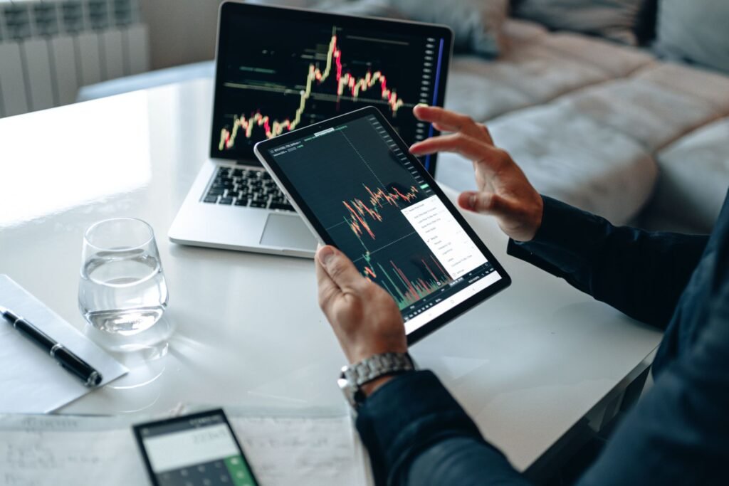 Close-up of a person using a tablet for online trading analysis with a laptop and glass of water nearby