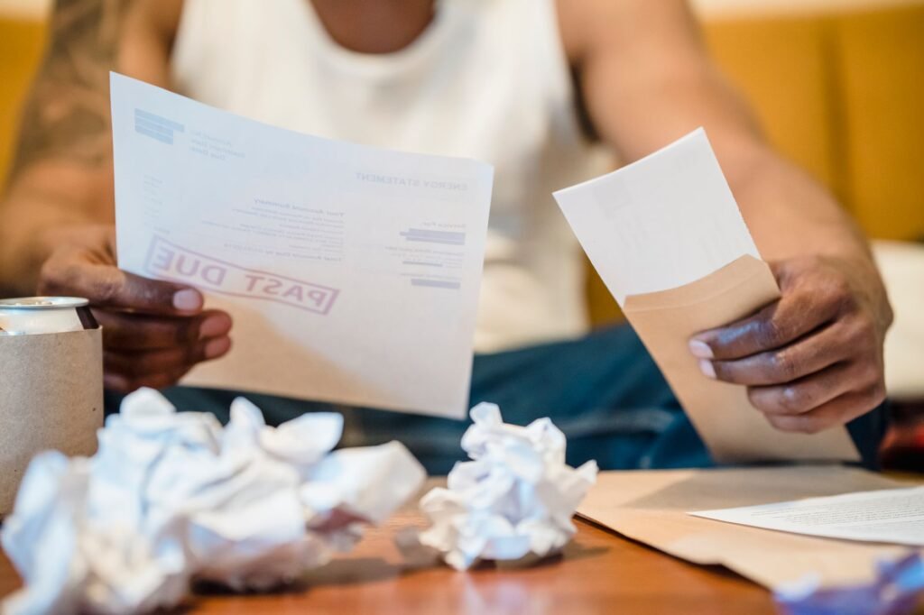 Man sitting indoors reviewing past due bills with crumpled papers on a coffee table