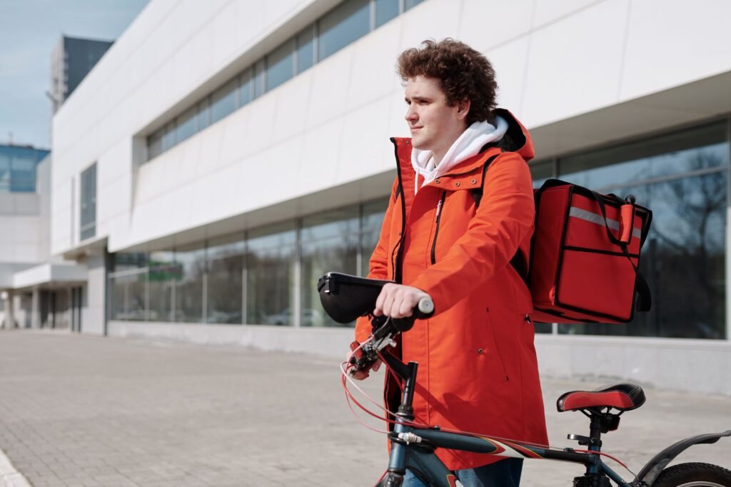 A bicycle delivery worker in a red jacket stands with a bike outdoors, ready for deliveries