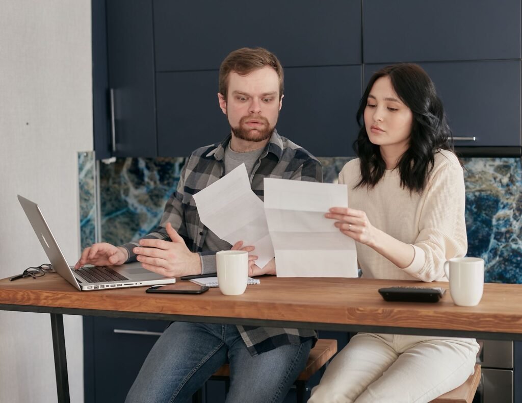 A couple sitting at a wooden table, reviewing documents with a laptop nearby, expressing concern