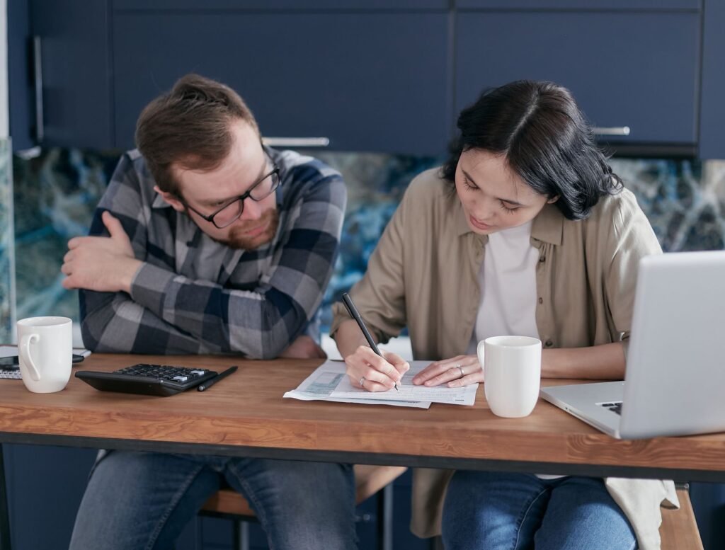 Adult couple working together on documents at a desk, with laptop and calculator nearby, in a home setting