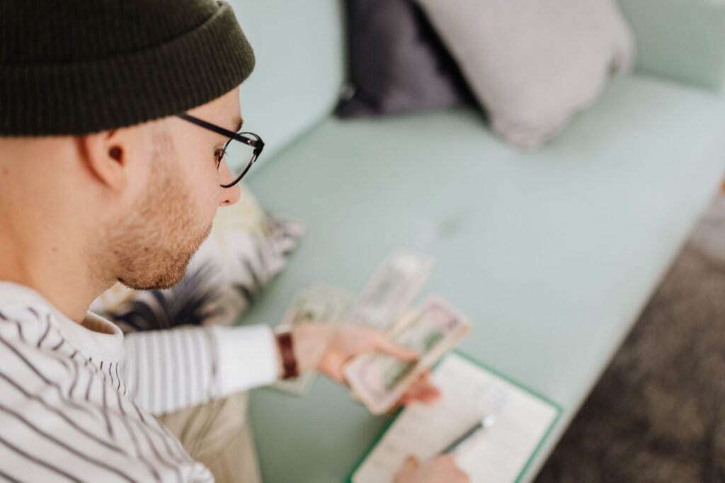 Adult man wearing glasses and beanie counting cash while budgeting indoors