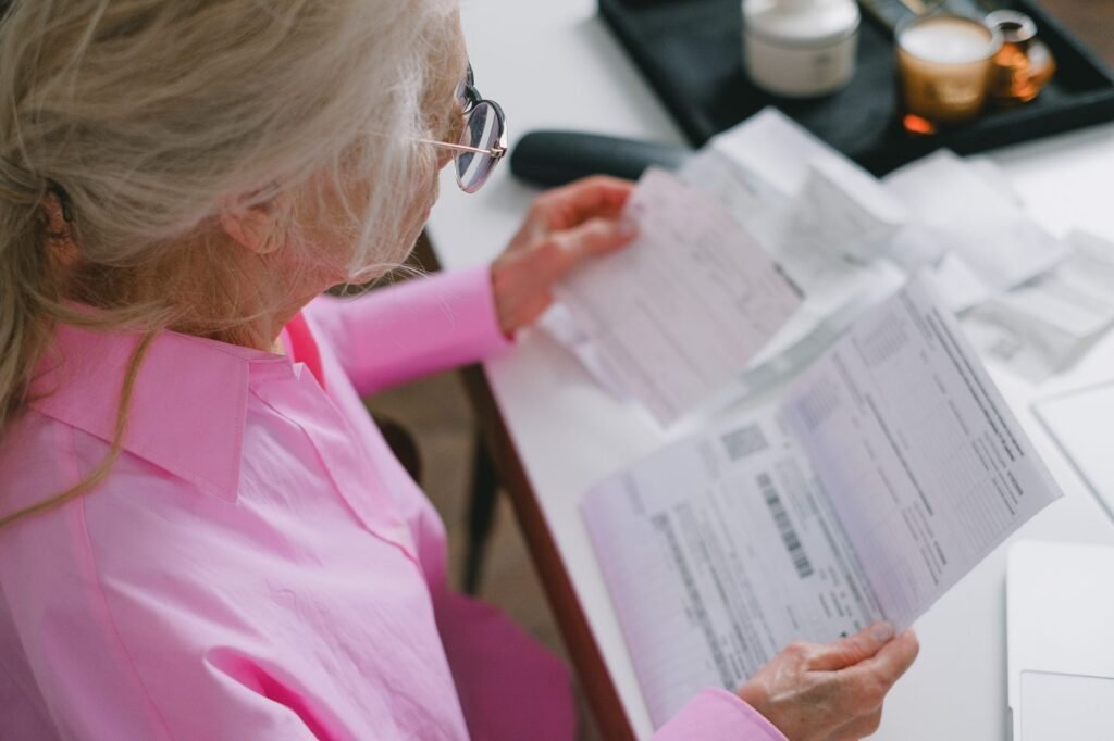 An elderly woman in glasses holds and reads important papers at a table indoors