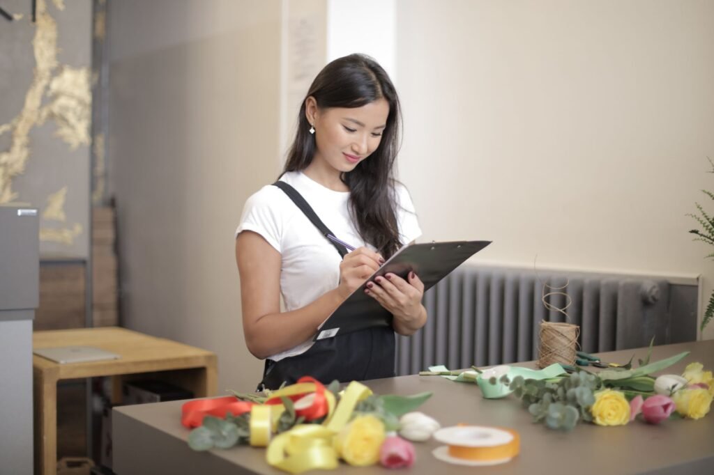 Asian female florist checking inventory in shop with flowers and ribbons on table