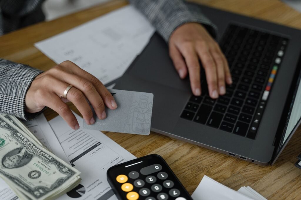 Close-up of a person using a credit card and laptop in a financial setting with cash and calculator